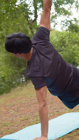 Vertical-Video-Of-Man-Wearing-Sports-Clothing-Doing-Yoga-Stretches-On-Mat-In-Forest-By-Lake-Or-River-Enjoying-Peace-And-Beauty-Of-Nature-Shot-In-Real-Time